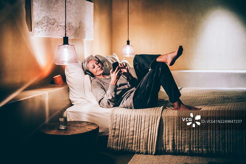 在家中的惬意时光：一位女士戴着耳机，一边听音乐一边使用智能手机，她舒适地躺在柔软的床上，在温暖舒适的客厅里享受着悠闲的个人时光。图片素材