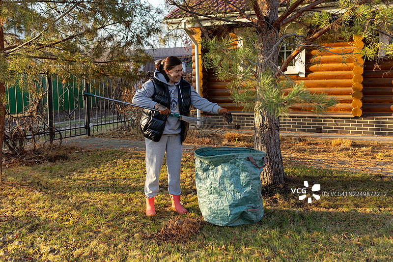秋日午后，一名女子在原木小屋旁清理院子，收集落叶，身旁放着一个大大的绿色袋子。图片素材