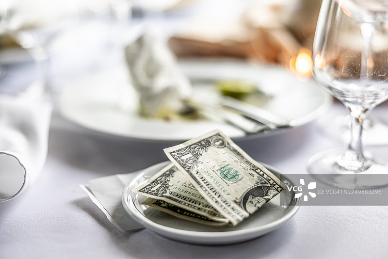 在餐厅的桌子上，一个小小的白盘子里放着折叠的美元纸币，这是给服务员的小费。背景中可以看到餐厅的餐具摆设，包括盘子里吃剩的食物。图片素材