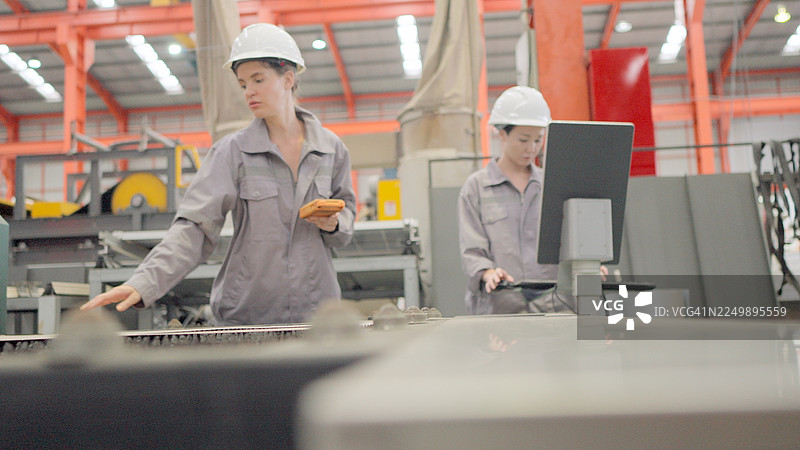 训练有素、经验丰富的生产线工人在工厂的制造和生产控制系统中，穿着安全制服在金属加工线上工作，将原材料板材装入机器进行成型。主管们在监控生产线。图片素材