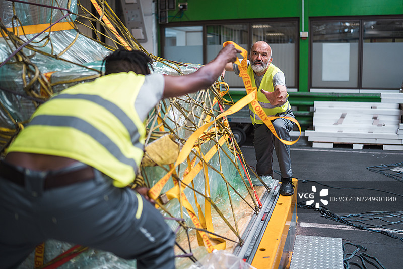 年轻的黑人和成熟的高加索男子在机场仓库穿着安全背心固定货物图片素材