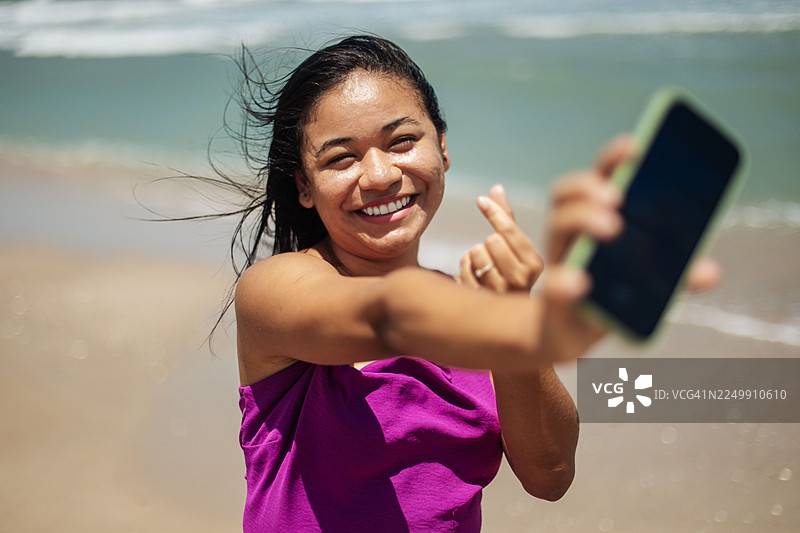 快乐的年轻巴西女子在海滩上自拍，并做出手指爱心动作图片素材
