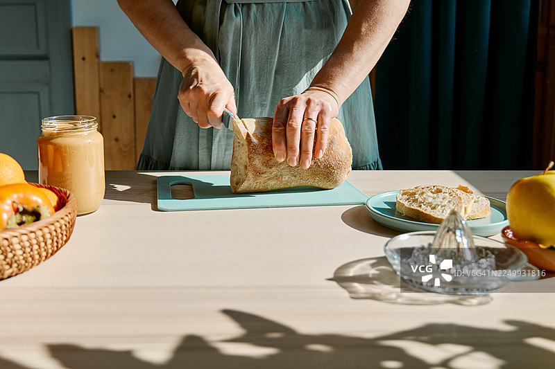 一位看不清面容的女性正在制作健康的早餐或早午餐，她在一块砧板上切着面包，旁边还有面包和花生酱。图片素材