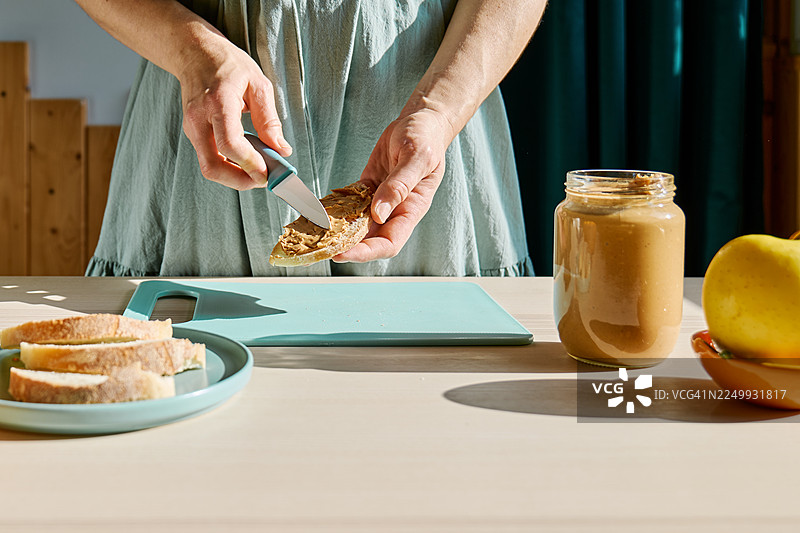 一位看不清面容的女性正在制作健康的早餐或早午餐，她在面包片上涂抹花生酱。图片素材