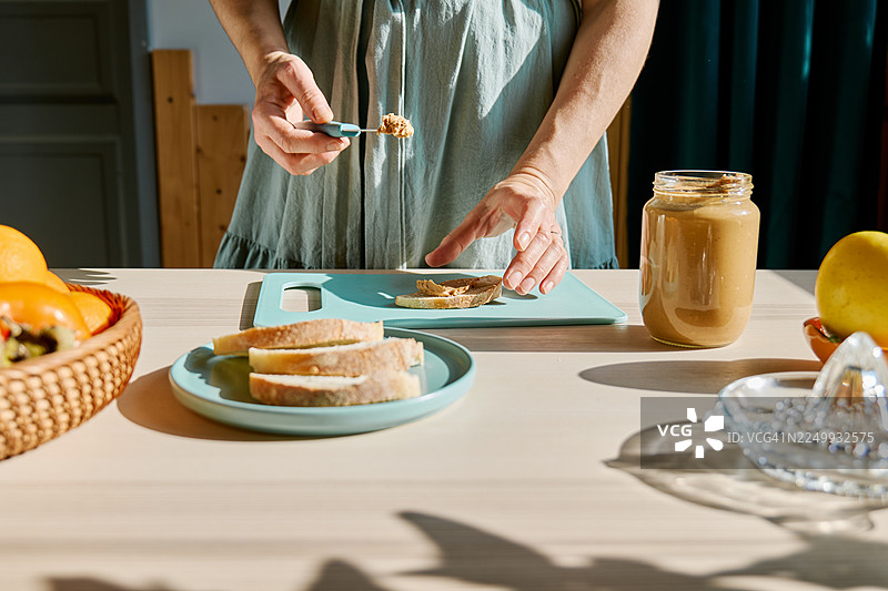 无法辨认的女性正在制作健康的早餐或早午餐，在吐司上涂抹花生酱。图片素材