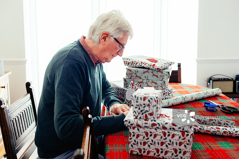 一位年长的男士在家中写圣诞贺卡，周围摆满了包装好的礼物。图片素材