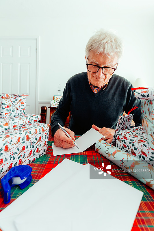 一位年长的男士在家中写圣诞贺卡，周围堆满了包装好的礼物图片素材