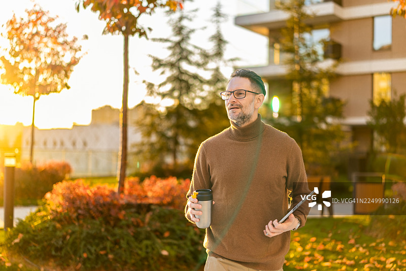 英俊的中年男子在秋季的城市里，手持平板电脑，眺望远方，享受咖啡休息时光，展现成功与未来愿景。图片素材