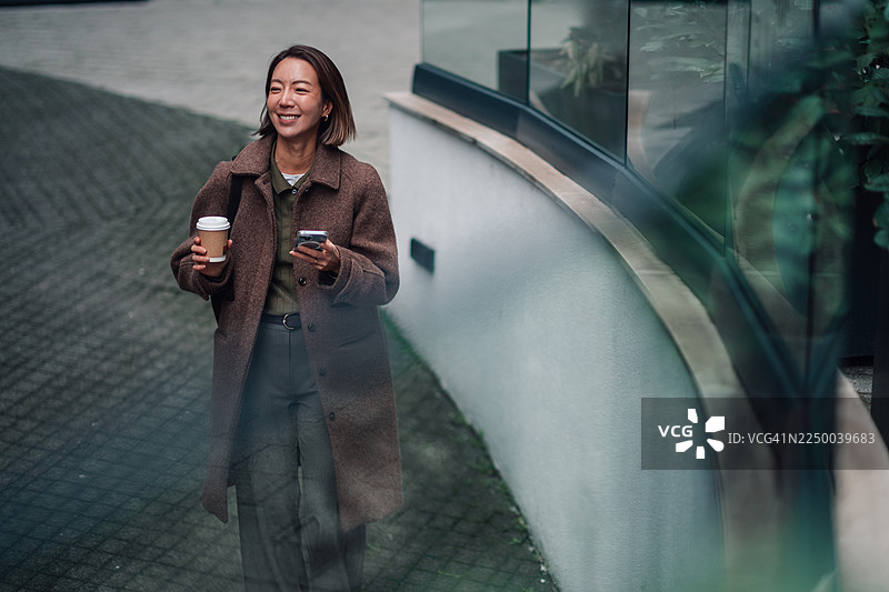 快乐的亚洲女性在城市街道上边走边使用手机喝咖啡图片素材
