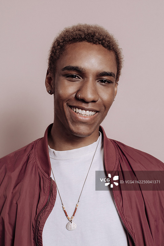 身穿酒红色夹克、佩戴珠串项链的年轻男子，面带微笑，肖像照，背景为柔和的粉色图片素材