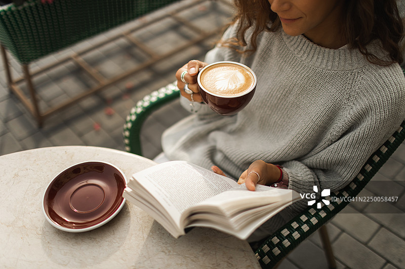 舒适的户外咖啡时光：身穿灰色毛衣的女子正在阅读，手边的拿铁咖啡有着拉花图案图片素材