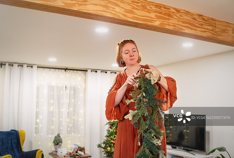 一位中年红发女性正在为冬季庆典布置节日绿花环，她在客厅里摆放着假期的装饰品。图片素材