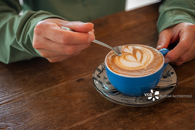 俯视角度拍摄，一个人在喝拿铁咖啡前，用勺子舀取咖啡表面的奶泡。图片素材