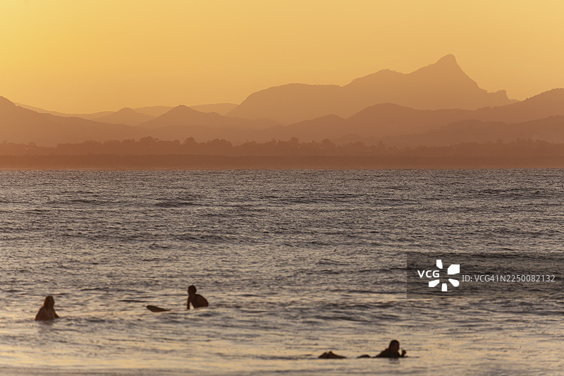 澳大利亚新南威尔士州拜伦湾瓦特戈斯海滩的红色日落，海中有冲浪者，背景是沃伦宾山（沃特格斯山）的冲浪场景。图片素材