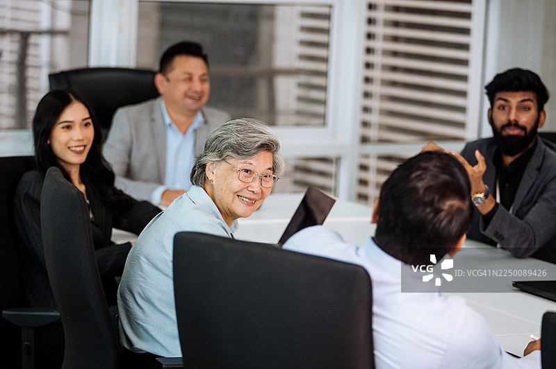 多元化的亚洲商务人士在现代办公室的办公桌旁协同工作，他们身着得体的商务服装，并使用笔记本电脑和数码平板等科技产品。图片素材