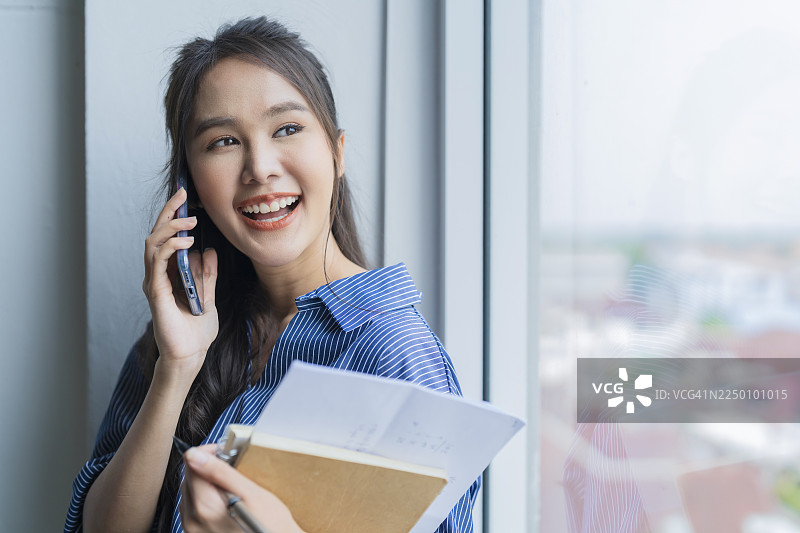 一位自信的女商人手持平板电脑，停下来回忆公司重要细节，随后她带着温暖的微笑，拎着文件板和智能手机，轻松自如地穿过现代办公室，展现出专业、抱负和成功的沉着风范。图片素材