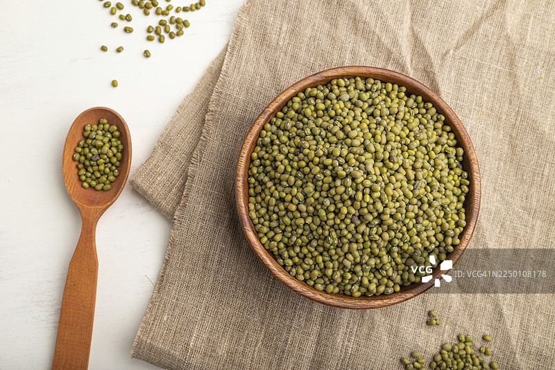 亚麻布料上摆放着装有生绿豆的木碗和木勺，背景为白色。俯视，平铺，特写图片素材