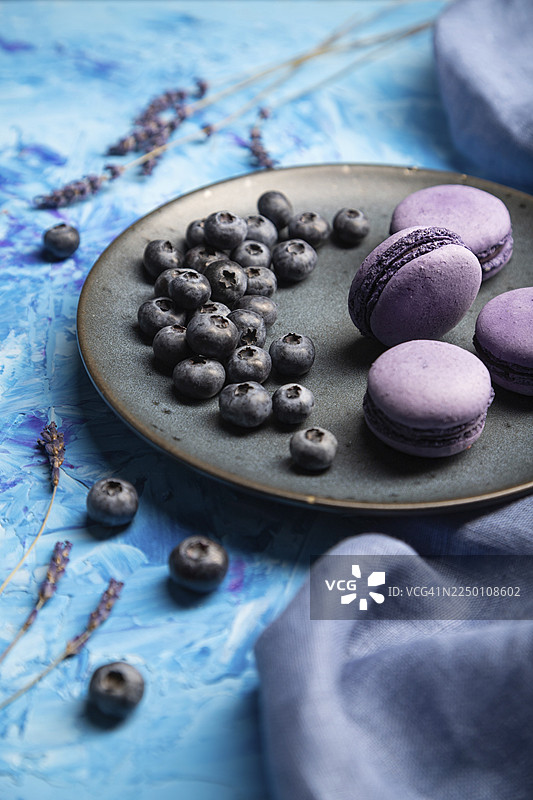 蓝色混凝土背景和蓝色纺织品上，陶瓷盘子里放着紫色的马卡龙或马卡龙蛋糕，上面点缀着蓝莓。侧视图，特写，选择性聚焦。图片素材