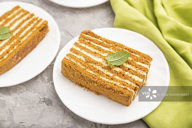 自制蜂蜜蛋糕搭配奶油和薄荷，旁边放着一杯咖啡，背景为灰色水泥地和绿色织物。侧视图，特写，选择性对焦。图片素材