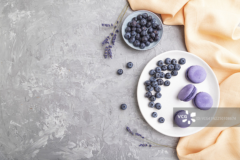 紫色的马卡龙配蓝莓，放在白瓷盘上，背景为灰色混凝土和橙色织物。俯视角度，有空白空间可供文字添加。图片素材