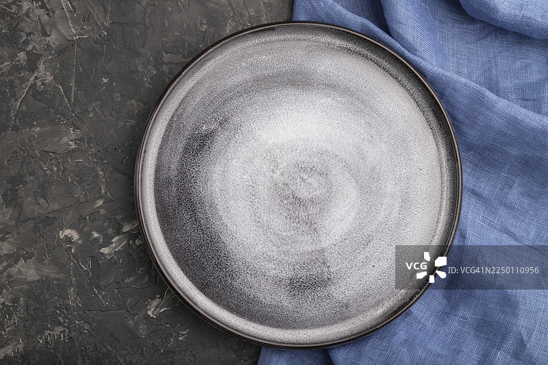 空置的灰色陶瓷盘放在黑色混凝土背景和蓝色亚麻纺织品上。俯视，特写，平铺图片素材