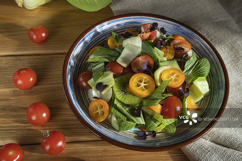 白菜、猕猴桃、番茄、金橘、微型蔬菜苗的素食沙拉，置于木质背景和亚麻纺织品上。强光，高对比度。侧视图，特写。图片素材