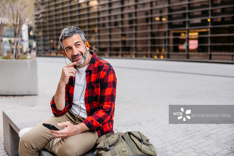 一位成熟的男性坐在巴塞罗那街头的长椅上，戴着有线耳机听音乐并使用手机。图片素材