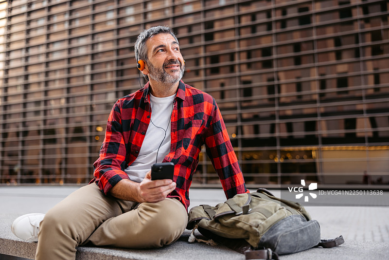 西班牙巴塞罗那街头长椅上，一位中年男子戴着有线耳机听音乐并使用手机。图片素材