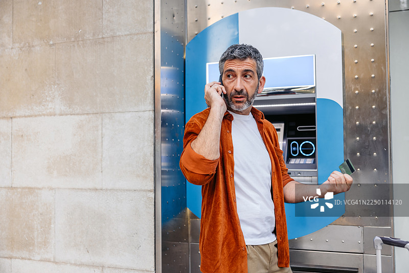 一名成熟的男子在巴塞罗那的ATM机前，一边拿着信用卡一边打电话。图片素材