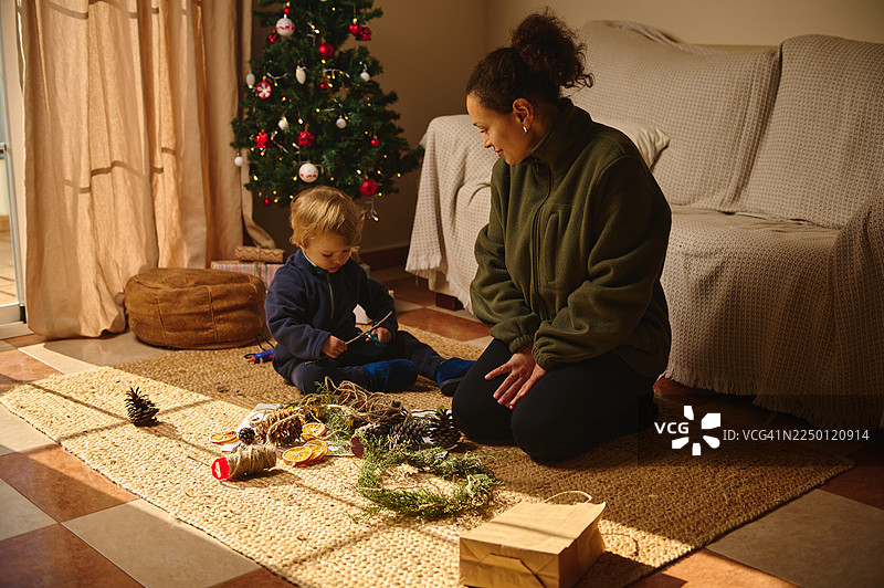 温馨的客厅里，妈妈和孩子在地毯上一起制作圣诞装饰品，旁边就是圣诞树。图片素材