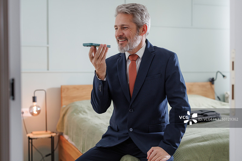 一位穿着西装的成熟男士，坐在床上，在现代卧室里打电话。图片素材