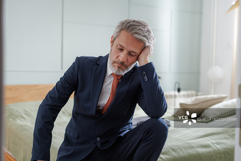 一位穿着西装的中年男子正坐在卧室的床上，感到疲惫。图片素材