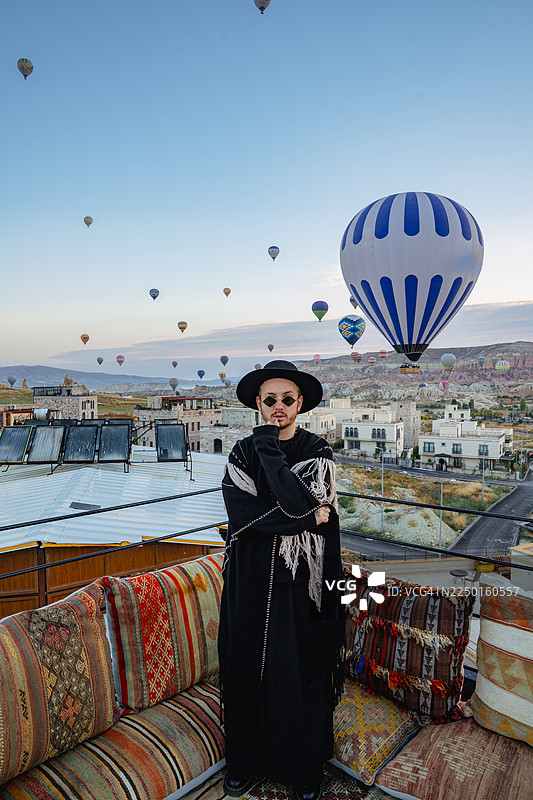 一位时尚的年轻男子身着潮流服饰，优雅地摆出造型，身后可见基里姆沙发和热气球，地点为土耳其卡帕多奇亚。图片素材