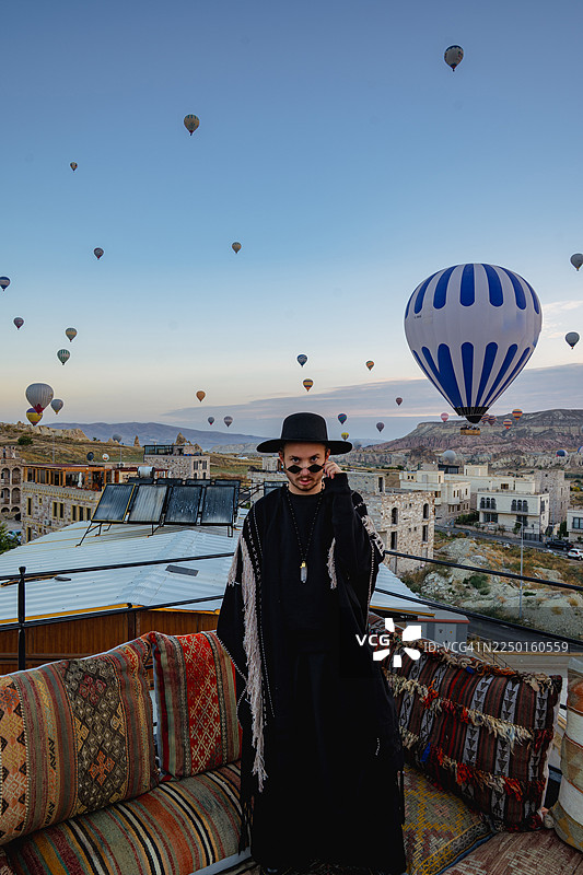 土耳其卡帕多奇亚，一位时尚的年轻男子站着摆姿势，背景中可以看到传统的基里姆沙发和热气球。图片素材