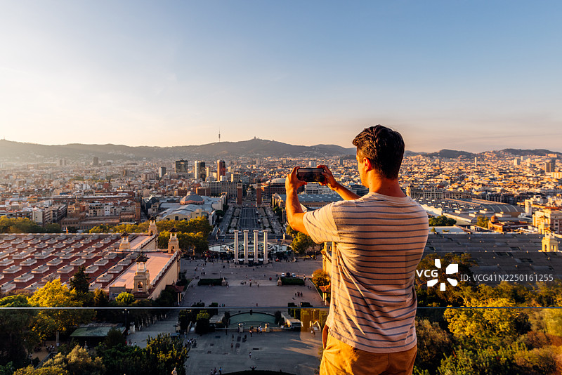 日落时分，一名男子在西班牙巴塞罗那用智能手机拍摄城市天际线，这是他的背影。图片素材
