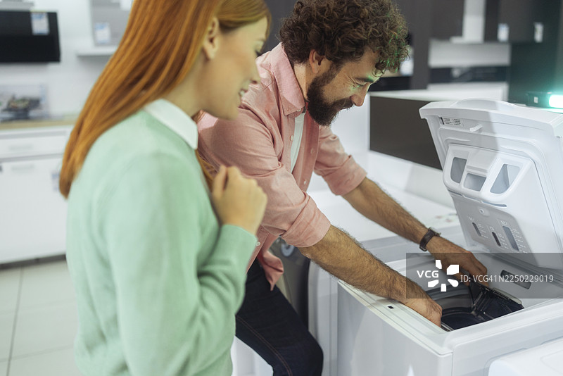 两名无忧无虑的顾客在一家电子产品商店，仔细查看一款新型顶开式洗衣机的内部。图片素材