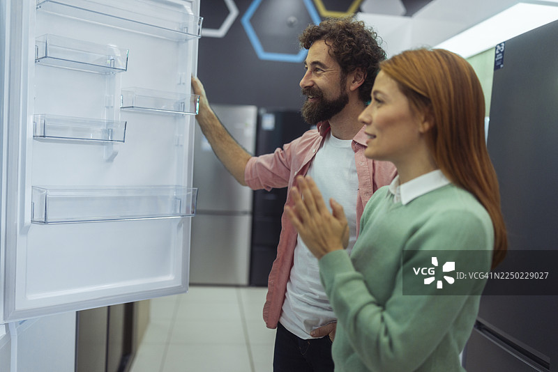 一家电子产品商店里，一男一女站在敞开的冰箱旁，正在考虑是否因为黑色星期五的额外折扣而购买它。图片素材
