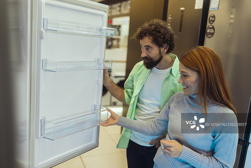 一对男女站在电子产品商店的敞开式冰箱旁，并考虑是否因为黑色星期五的额外折扣而购买它。图片素材