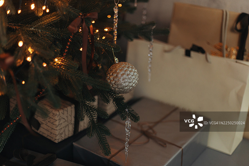 特写镜头，一棵圣诞树上装饰着彩灯、一个纹理金色挂饰和冰柱装饰品，背景是包装好的礼物和礼品袋。图片素材