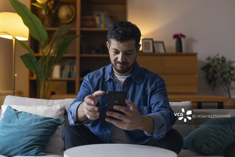 晚上，一个男人在家里的客厅里放松地躺在沙发上，浏览着智能手机，可能是在使用社交媒体、发短信或查看新闻。图片素材