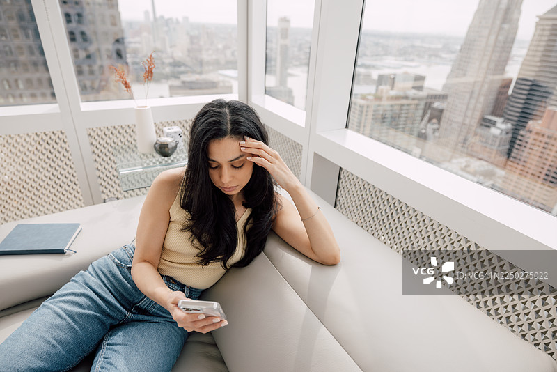 心情不佳的女人坐在沙发上，手托着头，背景是俯瞰纽约市中心的明亮公寓图片素材