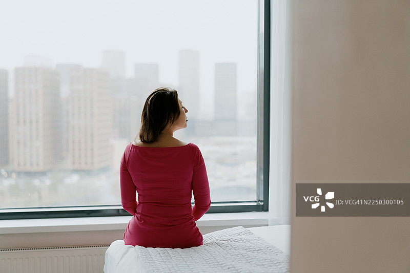 身穿粉色连衣裙的女子坐在床边，大窗户映照出宁静的城市天际线图片素材