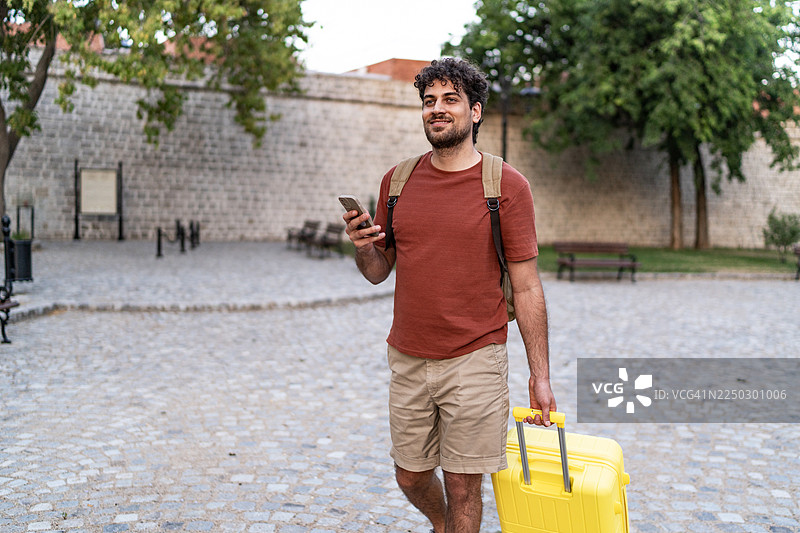 年轻男子拖着行李，手持智能手机，漫步在欧洲城市图片素材