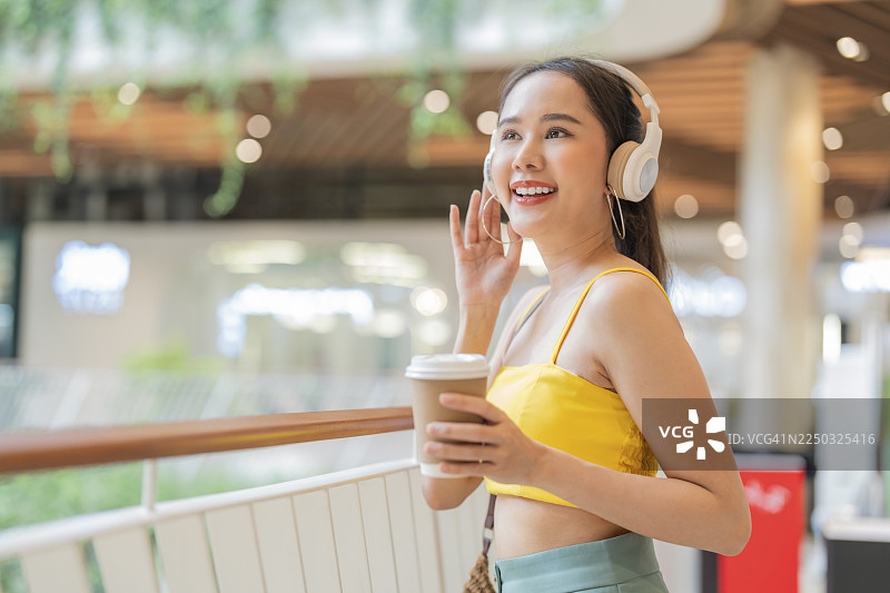 一位身着休闲夏装的亚洲女性，充满活力，在超市商场中漫步放松，她手持智能手机，一边选歌一边啜饮咖啡，享受着轻松自在的都市生活。图片素材