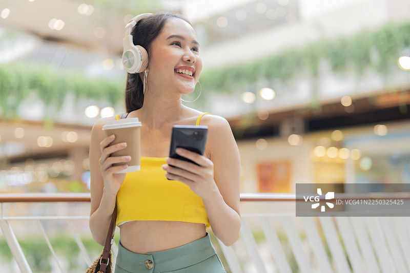 一位充满活力的亚洲女性，身着休闲夏装，在超市商场中漫步放松，她手持手机，一边选歌一边品尝咖啡，享受着轻松自在的都市生活。.图片素材
