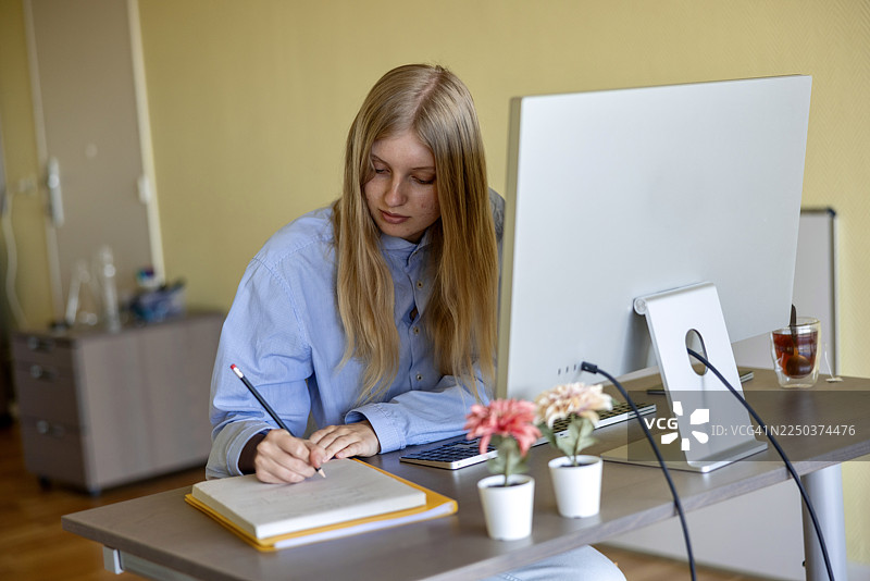 一位年轻的高加索女性在家中的办公桌前，坐在电脑显示器和盆栽植物旁边，正在笔记本上写笔记。图片素材