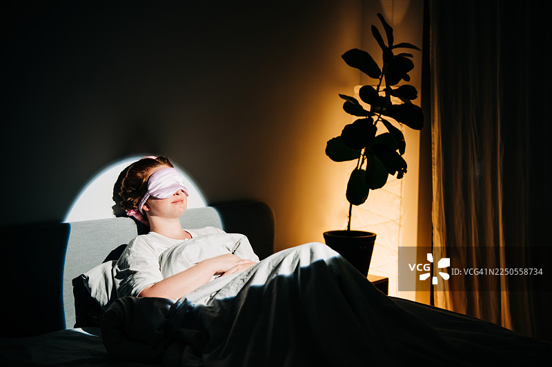 一个女人戴着眼罩躺在床上，卧室光线柔和，旁边有一盆室内植物。宁静的室内场景，表达了休息、自我关怀和宁静的夜生活方式。图片素材