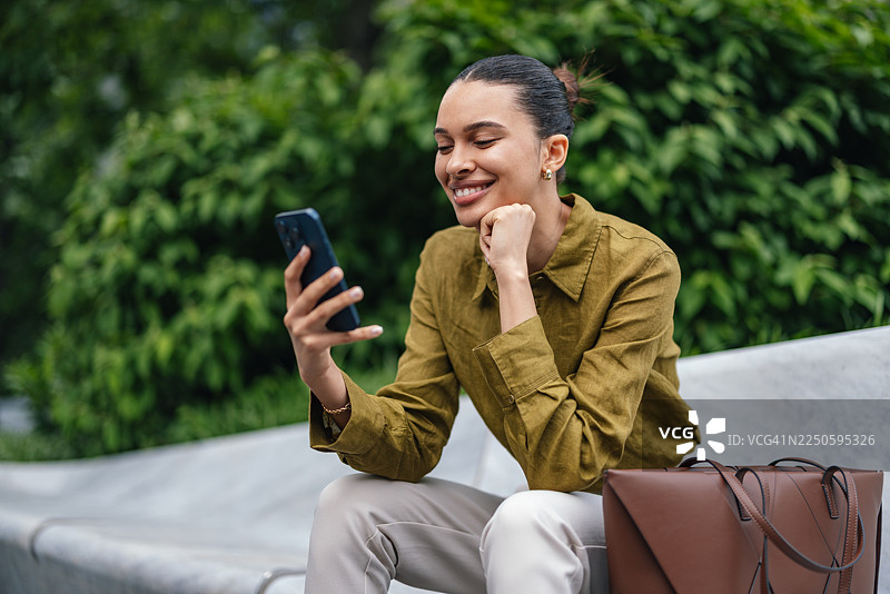 一位女士坐在城市公园的公园长椅上，微笑着使用智能手机，旁边放着一个棕色纸袋。图片素材