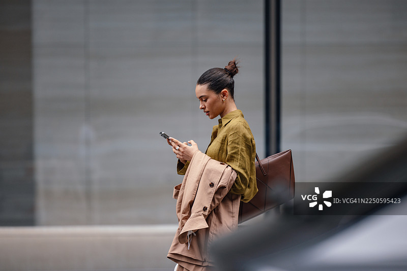 一位职业女性走在城市街道上，一边查看手机，一边将外套搭在胳膊上。图片素材