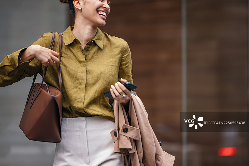 一位女商人在繁华的城市街道上，身披外套，手提包，面带灿烂的微笑，正在户外行走。图片素材